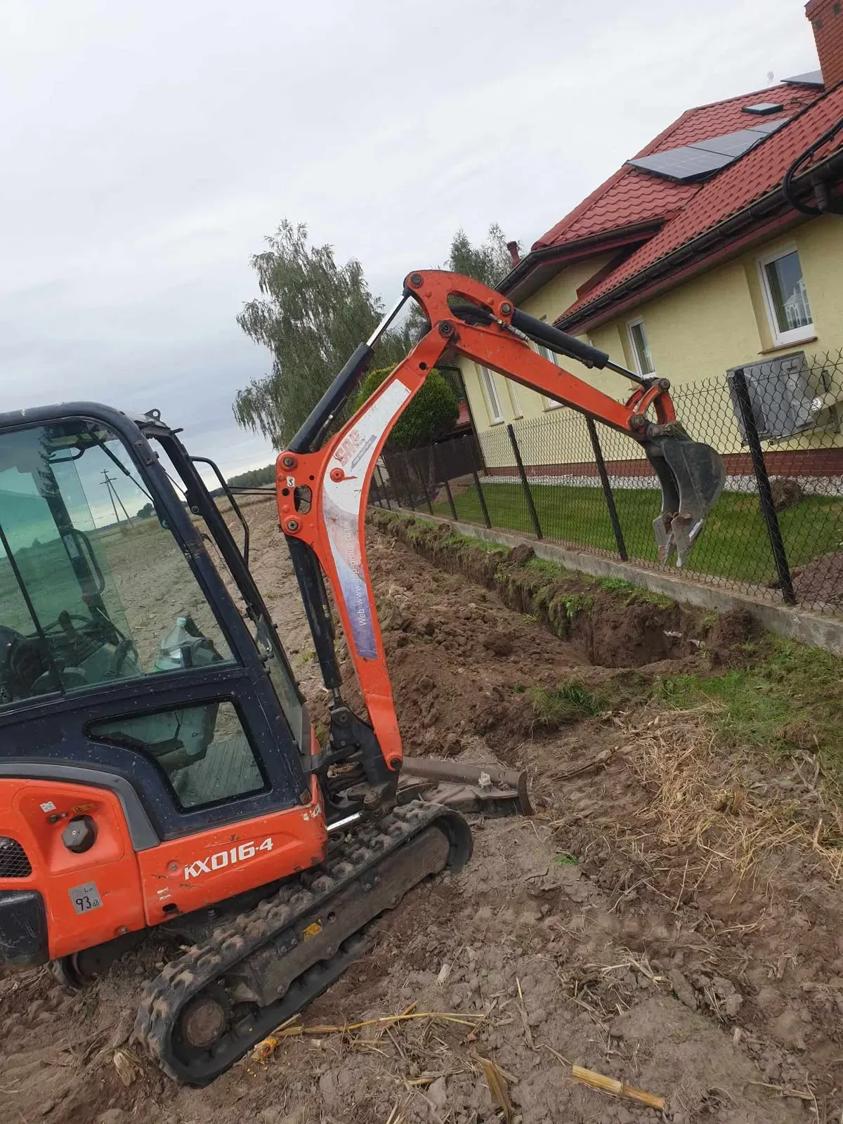 Pomarańczowa minikoparka kopiąca wykop pod przyłącze wody wzdłuż ogrodzenia domu