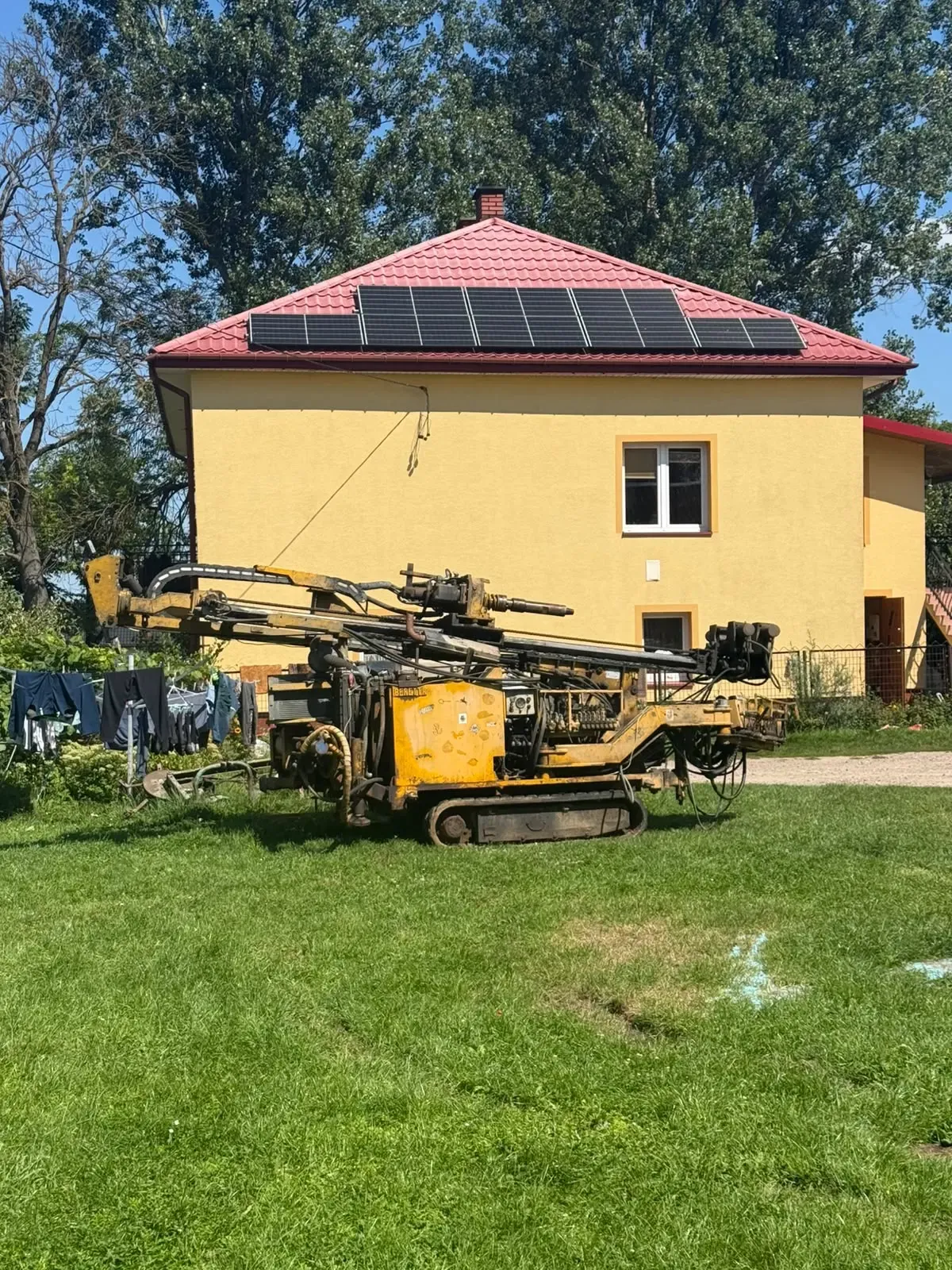 Żółta wiertnica gąsienicowa na trawniku przed domem z panelami fotowoltaicznymi na dachu